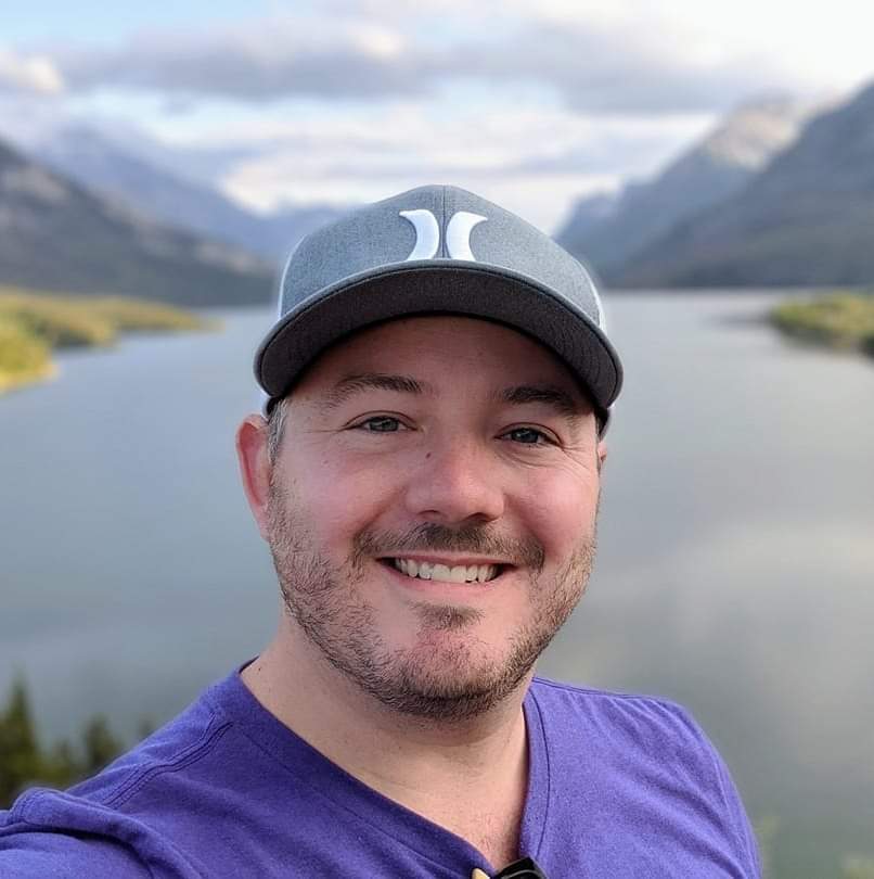 ACER alumnus Jason Weiler smiling in front of a scenic lake with mountains in the background, located in Waterton Lakes National Park, Alberta.